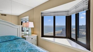 Un dormitorio con una cama grande, una mesita de noche, una lámpara y una ventana con vistas al océano y edificios.