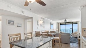 Amplia sala de estar con isla de cocina, mesa de comedor y sillas. Cuenta con ventiladores de techo, un ventanal con vistas y un balcón.