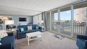 Ein modernes Wohnzimmer mit einem großen Schiebeglasfenster, das auf einen Balkon führt, einem an der Wand montierten Flachbildfernseher und Blick auf ein Hochhaus.