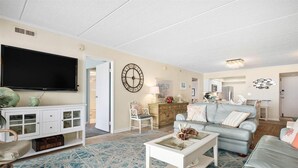 A living room with a television, white furniture, a blue patterned rug, and a large wall clock.