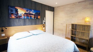 Une chambre d’hôtel moderne avec un grand lit, un tableau représentant un paysage urbain, une tête de lit en bois, une table de chevet avec une lampe et une étagère en bois.