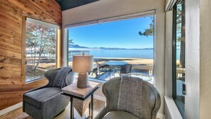 A room with a wooden wall, a large window with a view of a beach and water, two chairs, and a table with a lamp. - Stunning Lakefront Near Heavenly LLV611 (South Lake Tahoe)