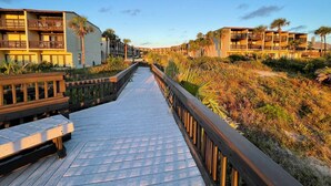 Un paseo marítimo de madera que conduce a un hotel frente a la playa, con varios balcones y vista al océano.