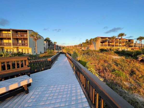 A wooden boardwalk leading to a beachfront hotel with multiple balconies and a view of the ocean.