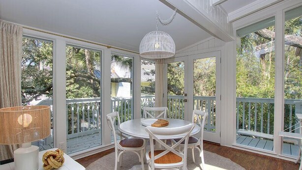A sunlit room with a round table and white chairs, large windows, and a white ceiling fan. - "Driftwood" in S. Forest Beach! (Hilton Head Island)
