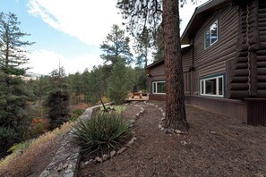 A log cabin with a deck, surrounded by dense forest and a stone pathway.