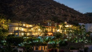 Un edificio de varios pisos con balcones y un jardín, rodeado de un paisaje montañoso.