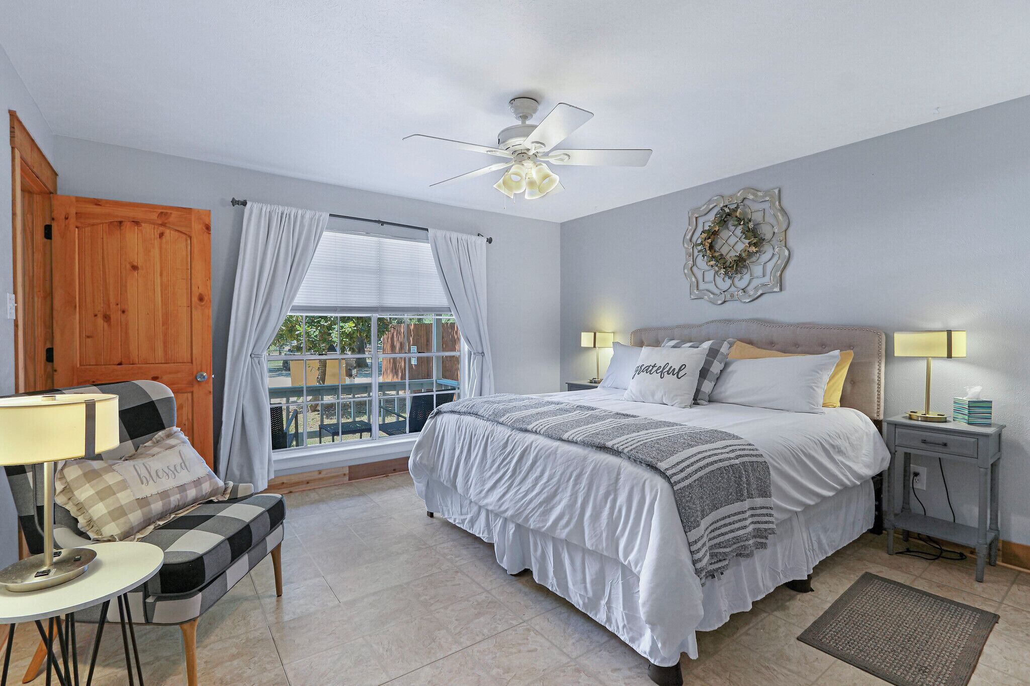 A bedroom with a bed, a chair, a nightstand, a lamp, a ceiling fan, and a window with curtains.