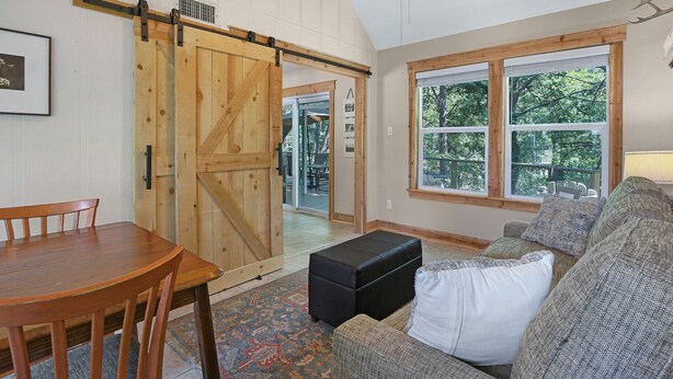 Una habitación acogedora con una mesa de madera, un sofá, una puerta corrediza tipo granero y ventanas amplias con vistas a árboles.