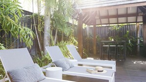 A sunlit outdoor seating area with white lounge chairs and a table, surrounded by lush tropical plants.