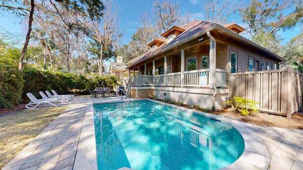 Une piscine entourée d’arbres, avec une terrasse en bois et des chaises longues, et une maison dotée d’un porche.