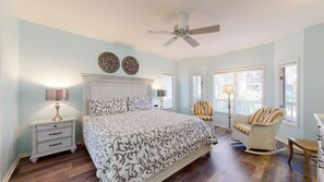 A bedroom with a large bed, a ceiling fan, two decorative wall pieces, a nightstand with a lamp, a rocking chair, and a window with blinds.