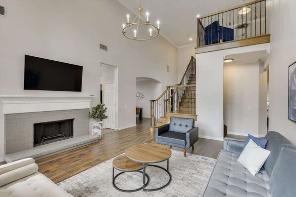 A spacious living room with a fireplace, a large flat-screen TV, a staircase, and a balcony.