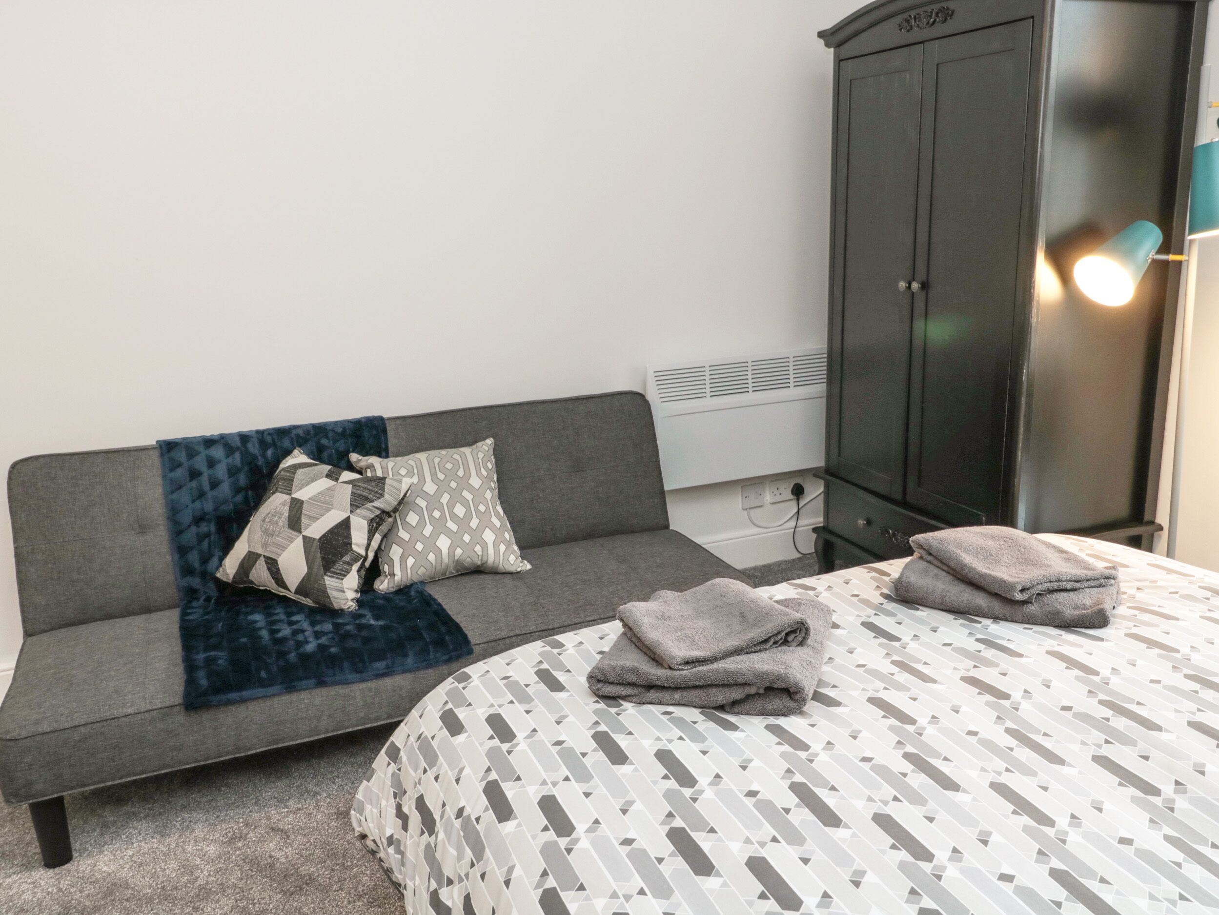 Een kamer met een grijze bank, een bed met een decoratieve spreidek en een donkere kast.