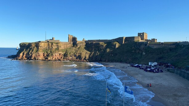 Zona costera con una fortificación histórica en un acantilado, una playa con sombrillas y una bandera.