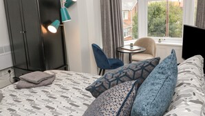 Un dormitorio con cama, armario, escritorio con silla, una mesita con planta y ventana que ofrece vistas a edificios y árboles.