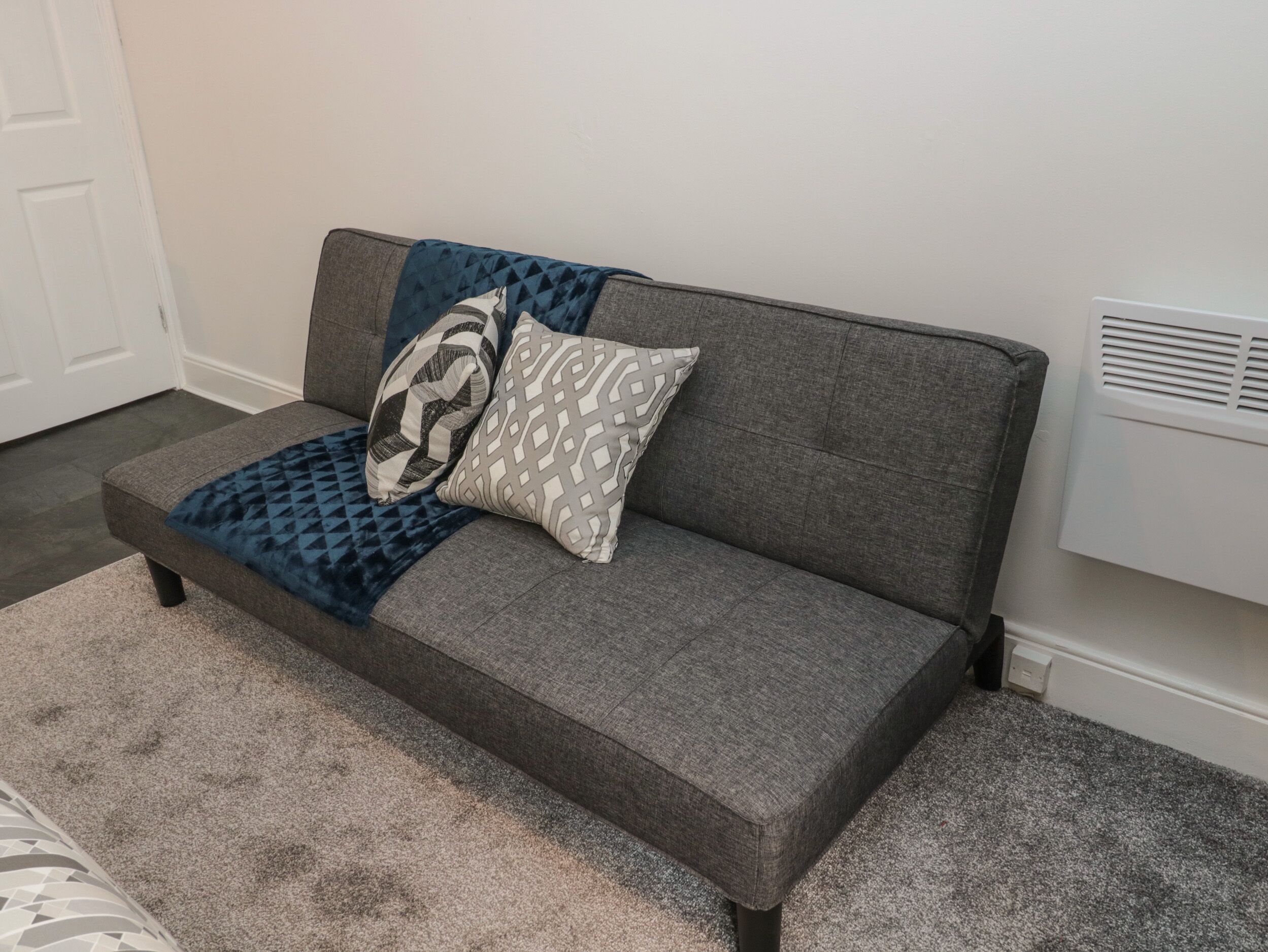 Un canapé gris agrémenté de coussins à motifs et recouvert d’un plaid bleu, placé sur un tapis gris dans une pièce dotée d’une porte blanche et d’un mur blanc.