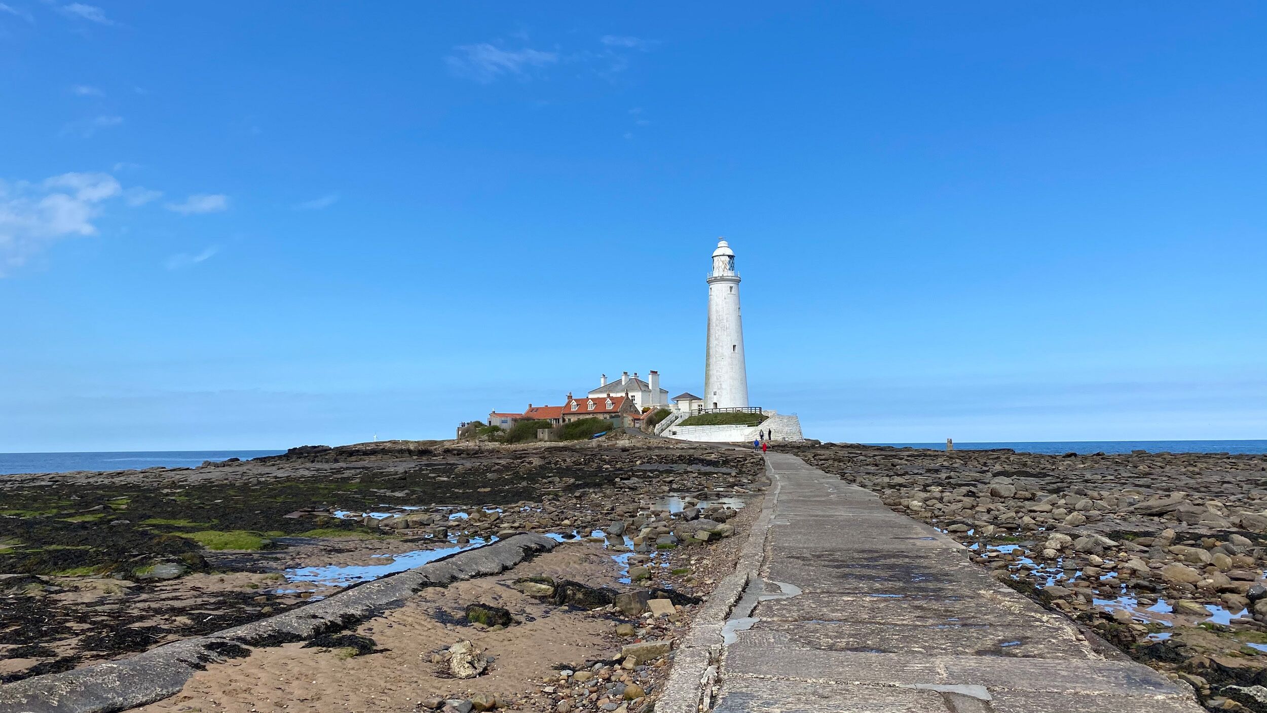 A coastal lighthouse with a concrete pathway leading to it, surrounded by rocky terrain and a clear sky.