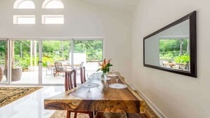A dining area with a wooden table, chairs, and a vase with flowers. There is a large window with a view of greenery outside.