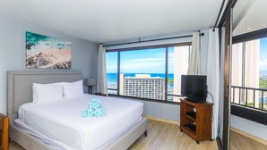 A hotel room with a large bed, a TV on a wooden stand, and a view of the ocean and buildings. - 2-Bedroom Ocean View, Perfect Location In Wakiki (Honolulu)