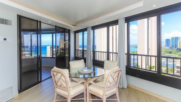 A modern room with a glass table and chairs, large windows with a city view, and a sliding glass door leading to a balcony. - 2-Bedroom Ocean View, Perfect Location In Wakiki (Honolulu)
