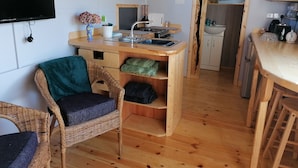 Ett kompakt kök med en bänkskiva i trä, en diskho och en mikrovågsugn. Där finns en flätstol med en kudde, ett litet bord och ett träskåp.