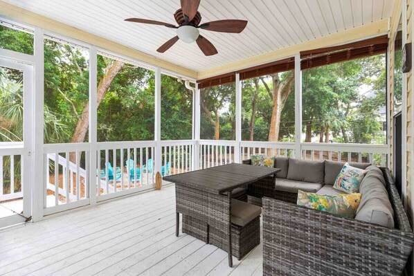 A covered porch with a table and seating, surrounded by white railings and trees.