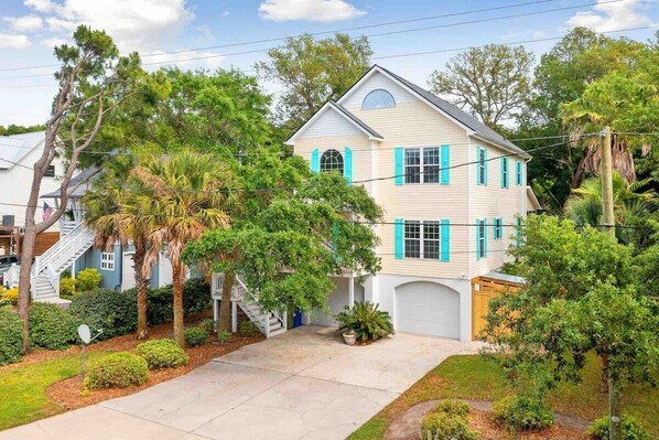 A two-story house with a garage, surrounded by lush greenery and a well-maintained lawn. - Folly Beach Gem: Walk to Beach, Restaurants & Nightlife - Happy Ours (Folly Beach)