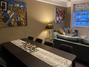 Un espace repas avec une longue table, des chaises et un élément décoratif au centre. On aperçoit un canapé avec des coussins et une lampe à l’arrière-plan.