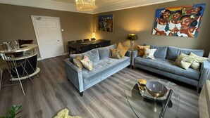 A modern living room with a grey sofa set, a glass coffee table, and a dining area with a chandelier. - Impeccable 4-bed Apartment in Bath (Bath)