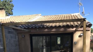 A covered outdoor seating area with two tables and stools, a tiled roof, and a sliding glass door leading to an interior space. - Maisonette with garden (Mornas)