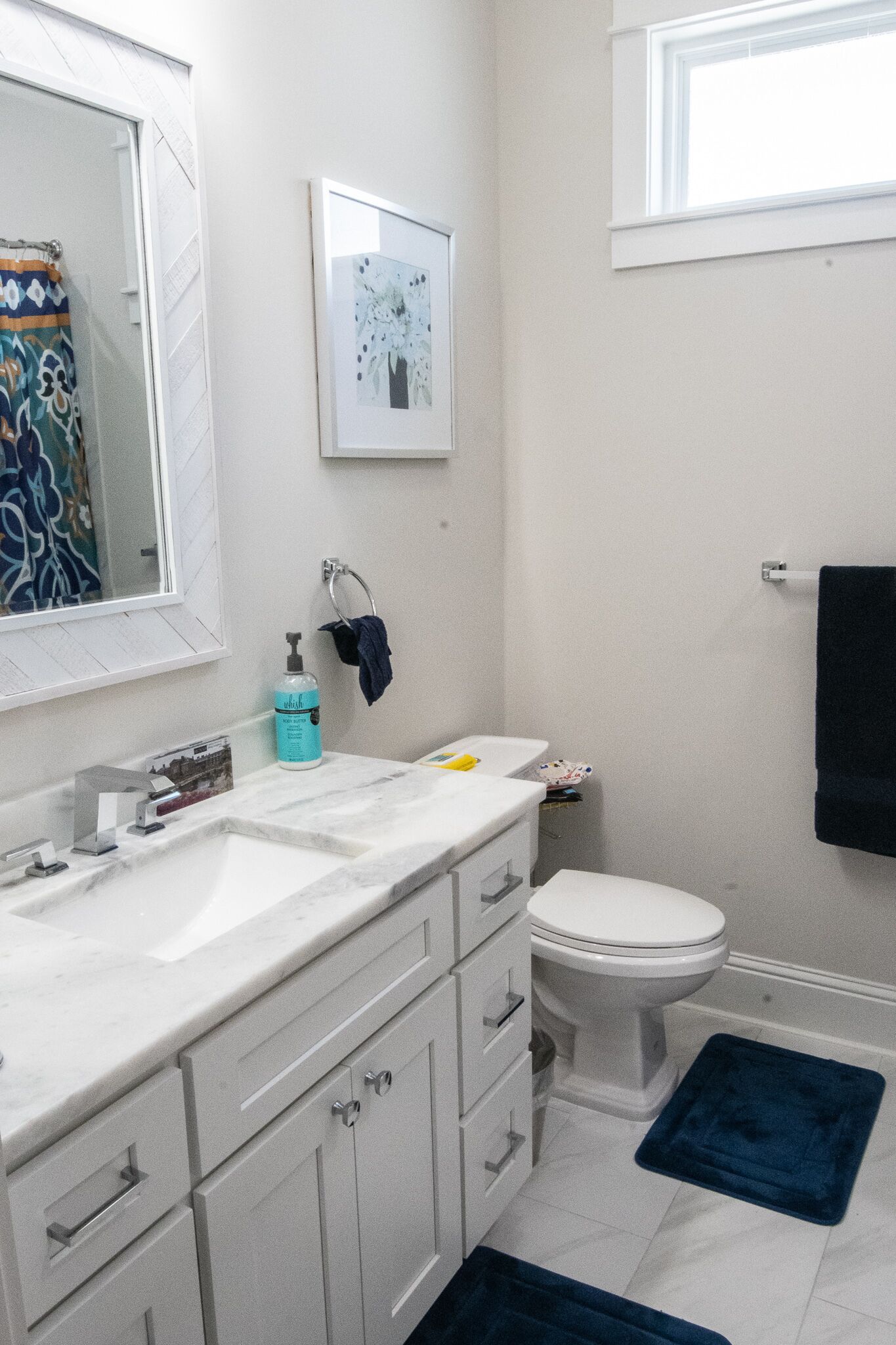 Baño con lavamanos blanco, espejo y retrete. Hay una alfombra de baño azul en el suelo.