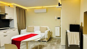 A compact hotel room with a kitchenette, a dining area with a red table runner, a white sofa, and a TV on a stand. - PANTHEON Maison, in the center of Rome + breakfast included (Roma)