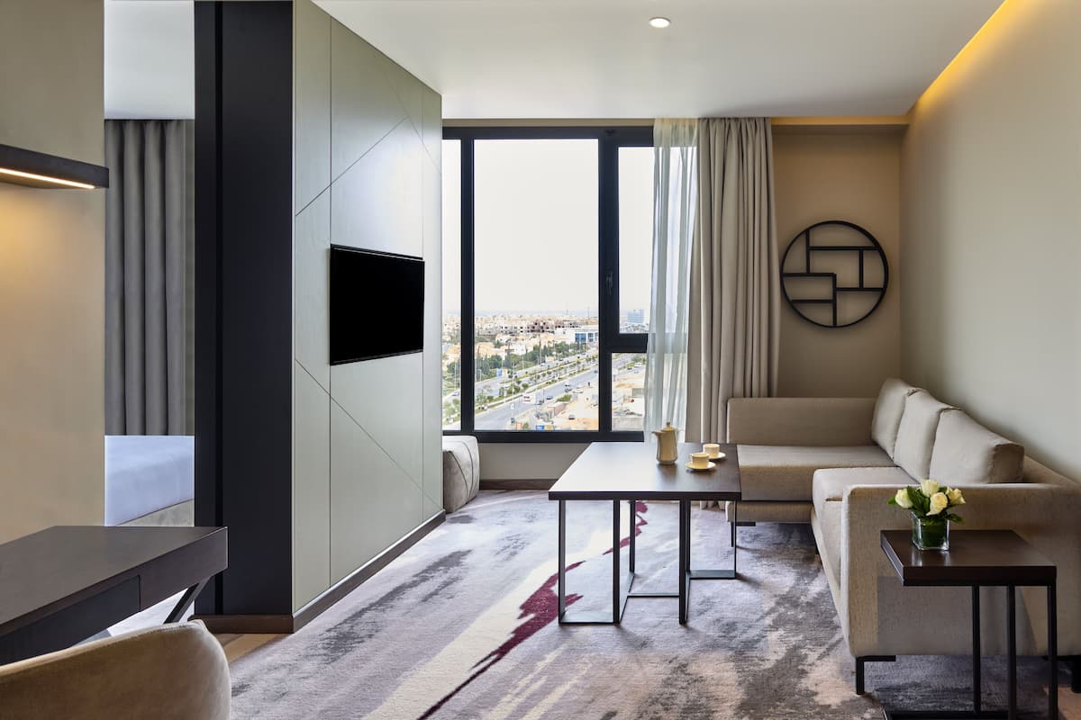 Une chambre d’hôtel moderne dotée d’une grande fenêtre, d’un téléviseur à écran plat, d’un canapé, d’une table basse et d’une petite table d’appoint sur laquelle est posé un vase de fleurs.