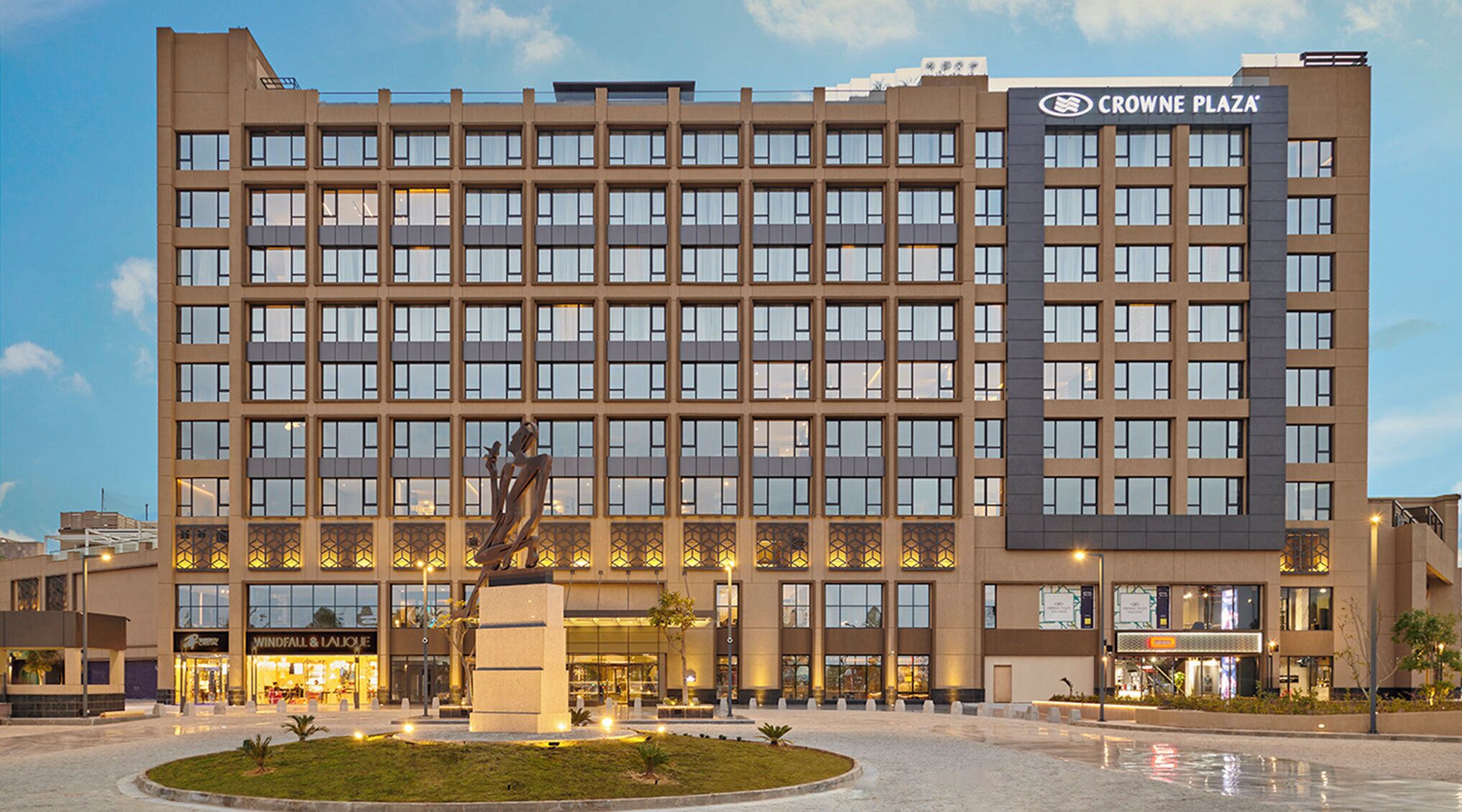 Un hotel de varios pisos con un llamativo letrero de Crown Plaza, un patio central con una escultura, y tiendas como Windfall & Lalique.