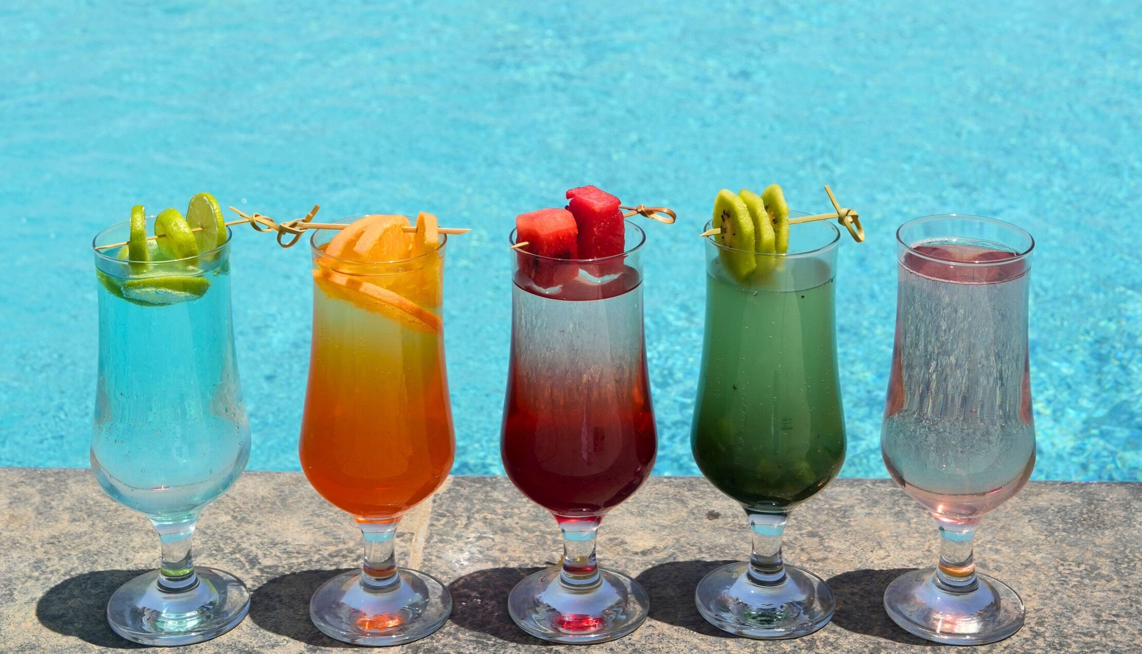 Cinq boissons colorées, décorées de morceaux de fruits, disposées sur une surface en pierre, près d’une piscine.