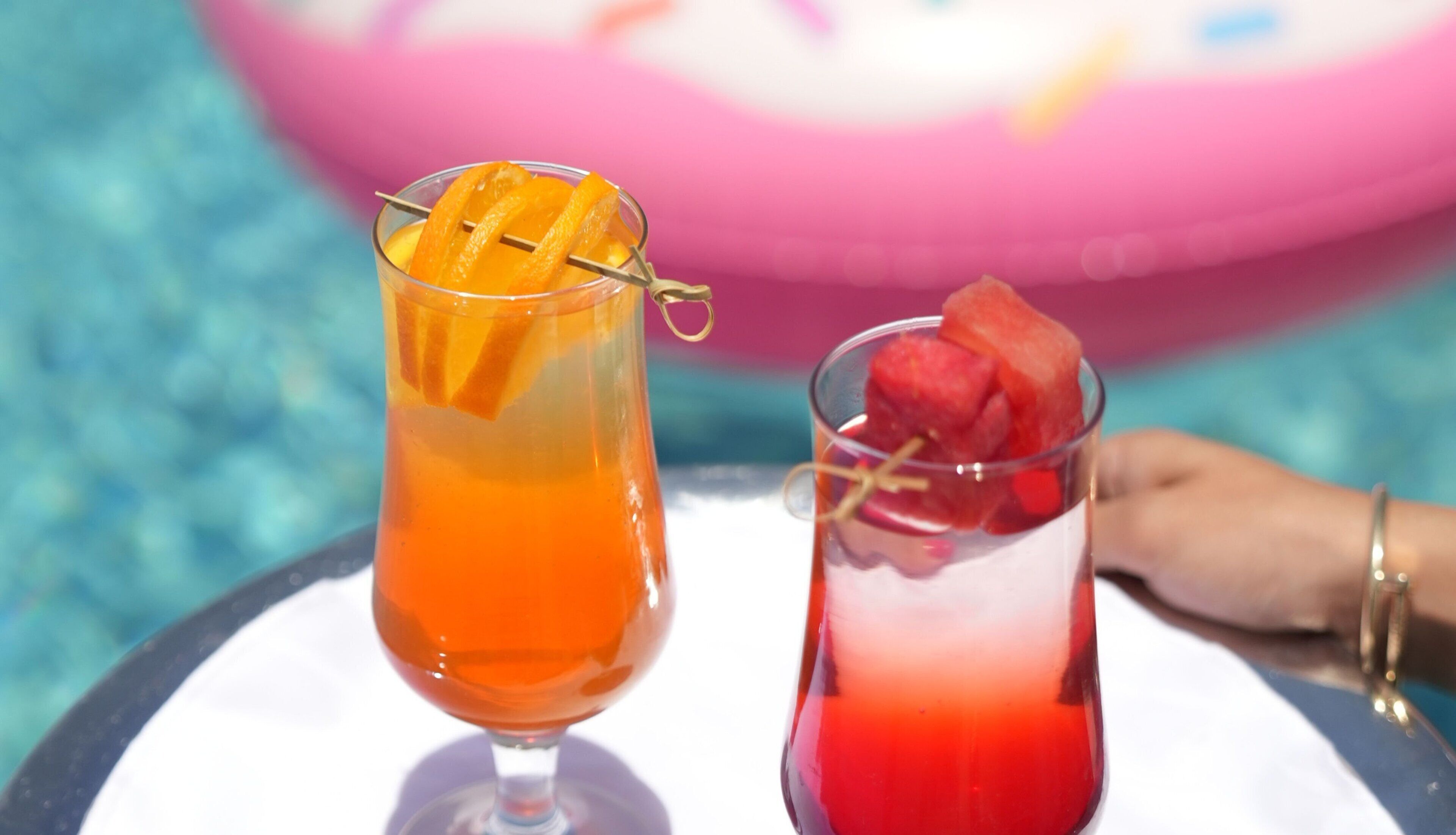 Dos bebidas en vasos de cristal con adornos de frutas, sobre una mesa con una piscina al fondo.