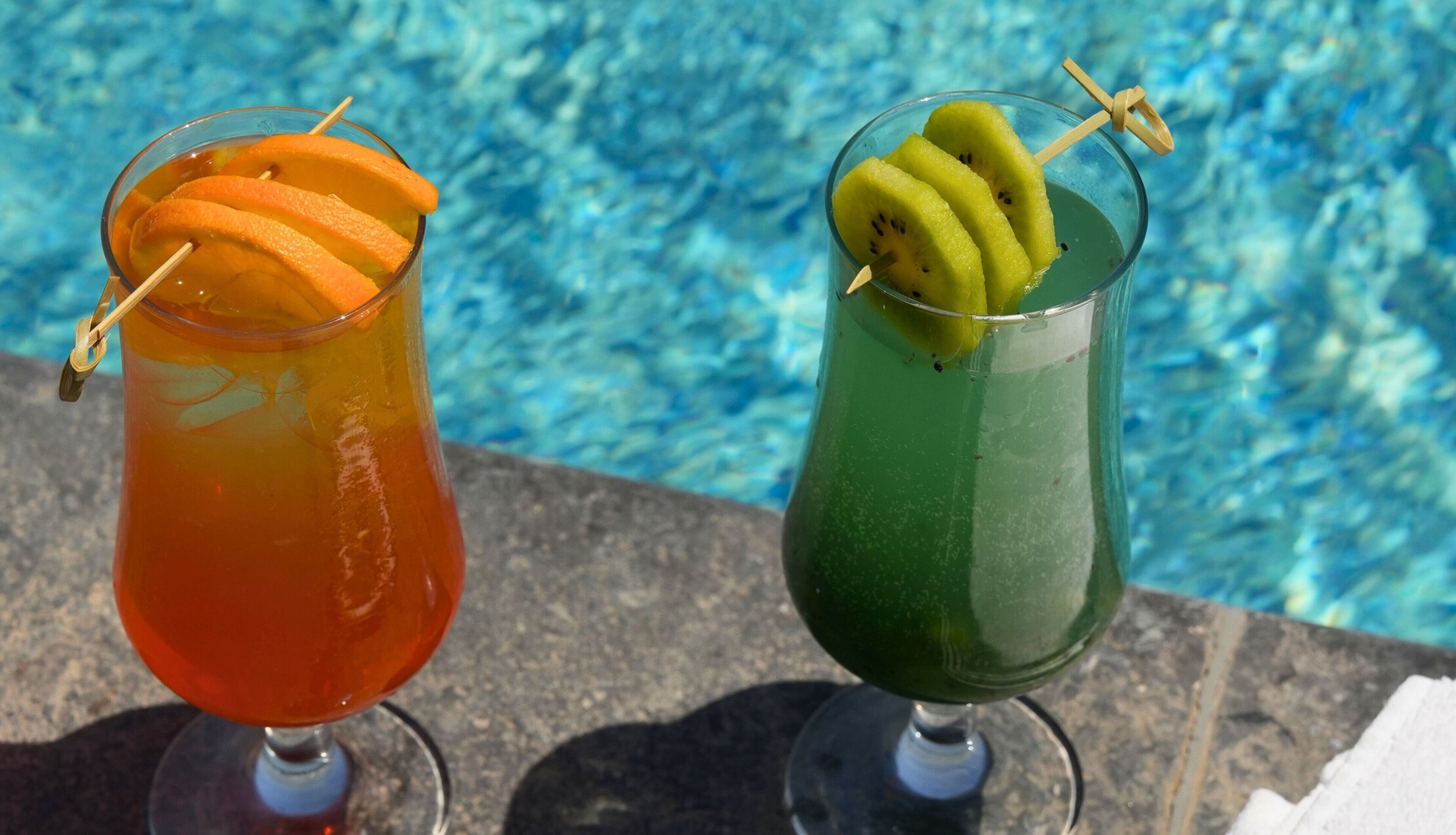 Two drinks with fruit garnishes by a pool.