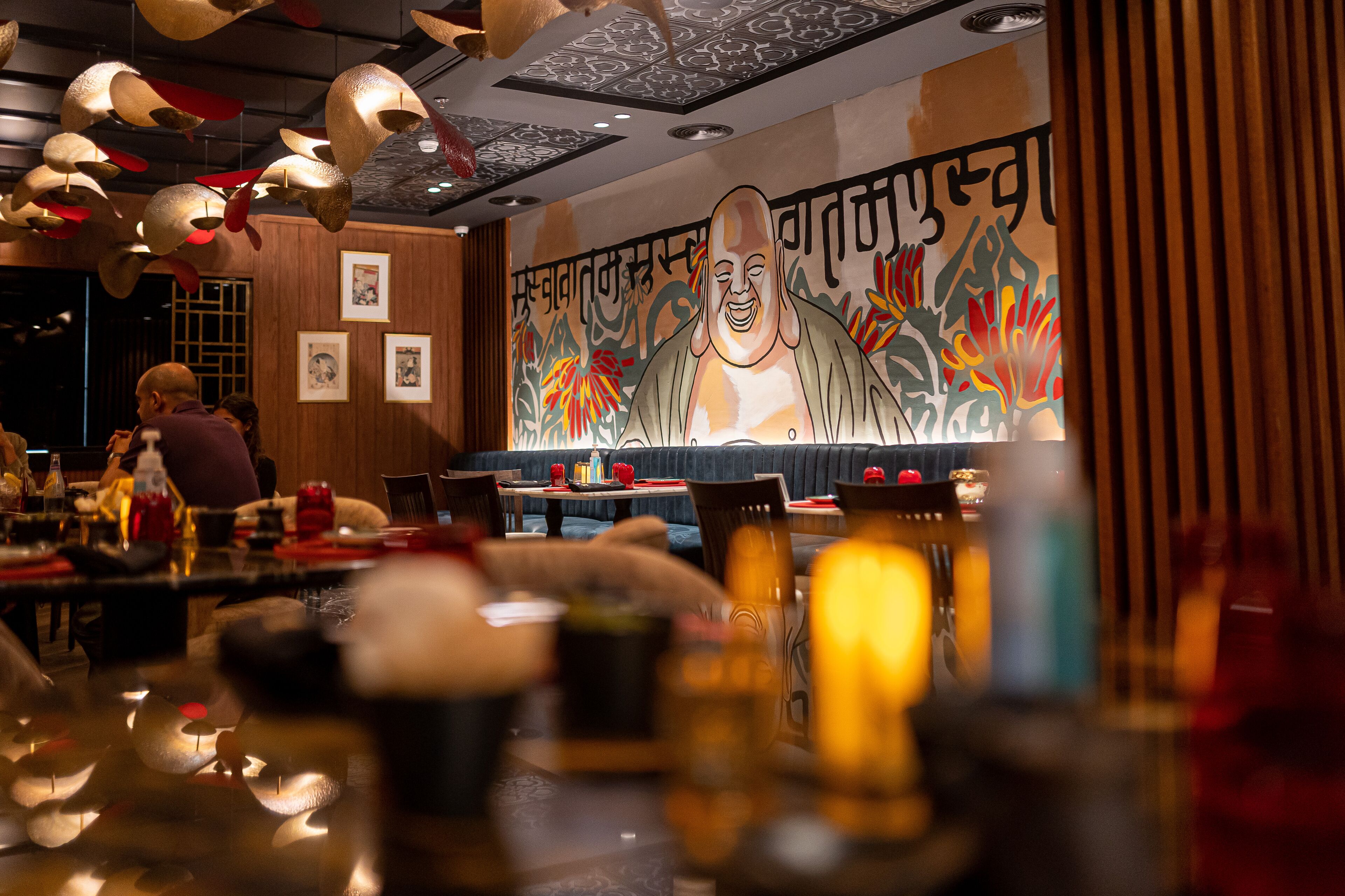 Restaurante com um mural de um Buda sorridente, paredes de painéis de madeira e luminárias pendentes.
