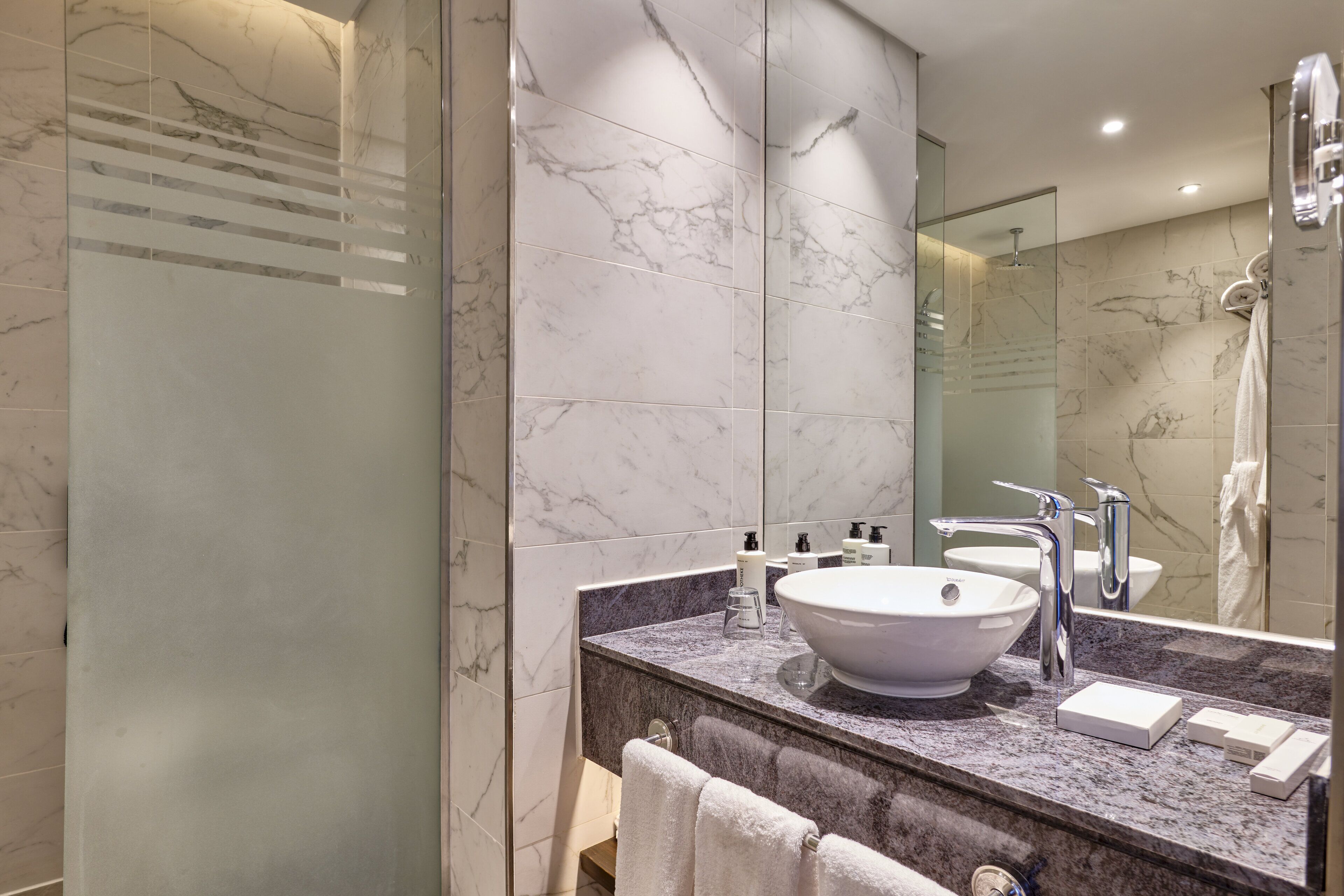 A modern bathroom with a large mirror, marble tiles, a white sink, and a shower area.