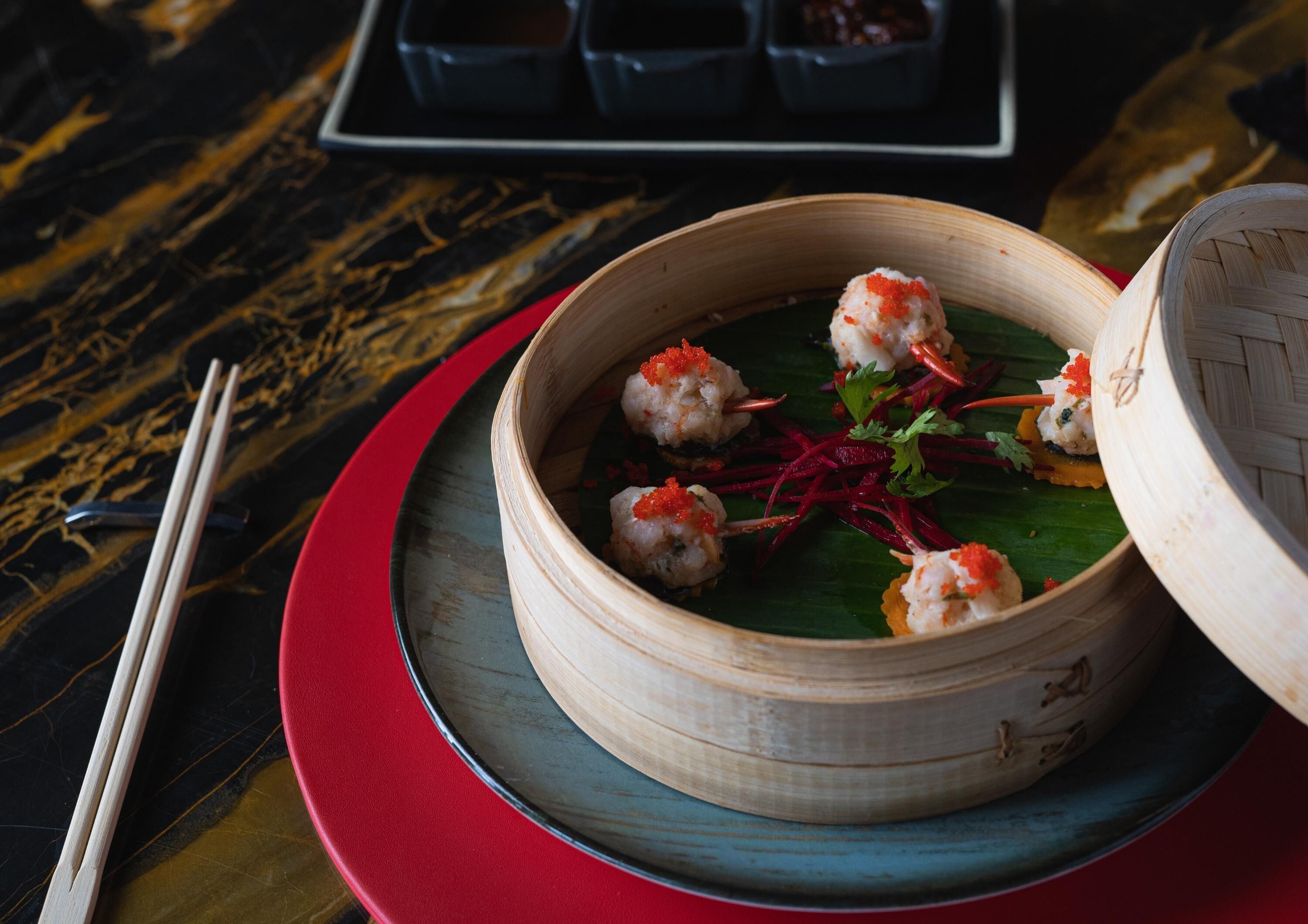 A bamboo steamer with dumplings on a red plate, accompanied by chopsticks and a small dish of sauces.