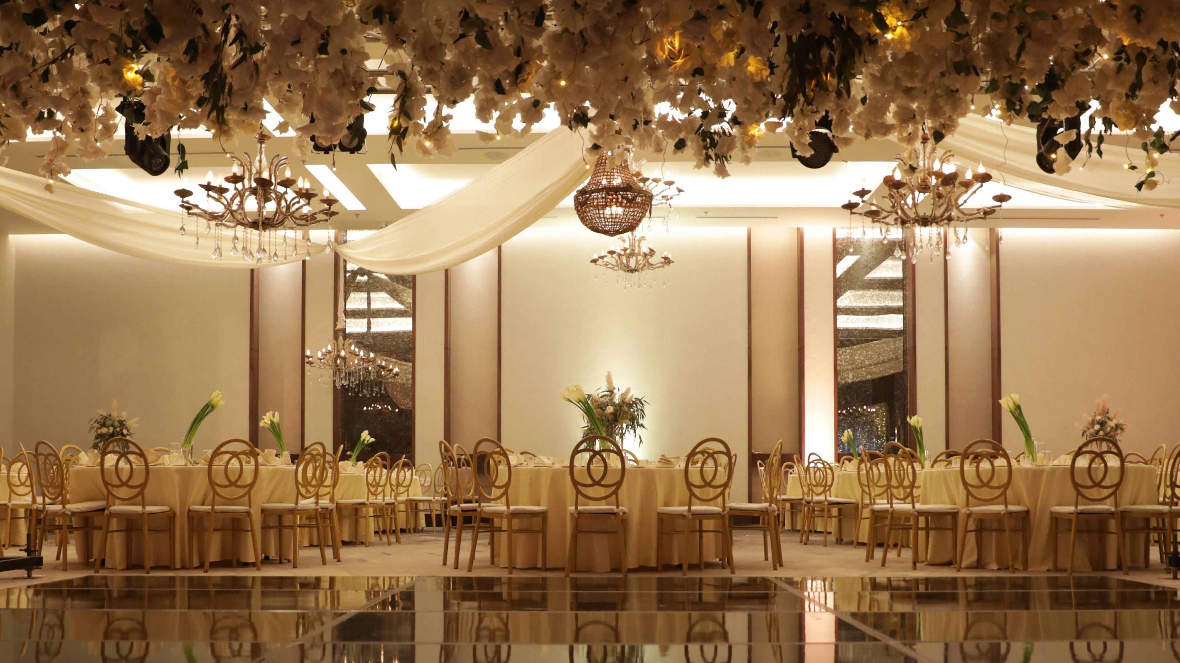 A banquet hall with round tables, chairs, chandeliers, and draped fabric.