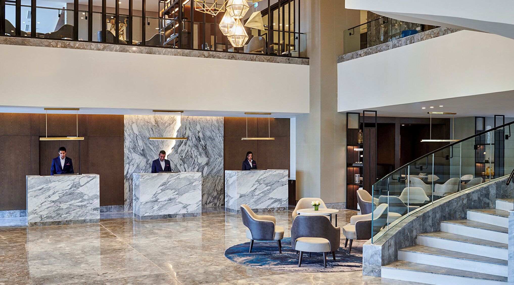 A hotel lobby with marble reception counters, a staircase, and modern lighting fixtures.