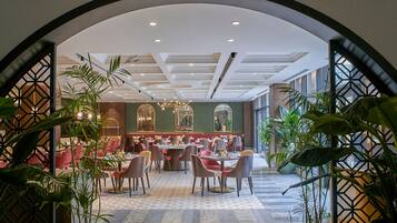 Un amplio comedor con múltiples mesas y sillas, techo alto con iluminación empotrada, espejos decorativos y plantas.