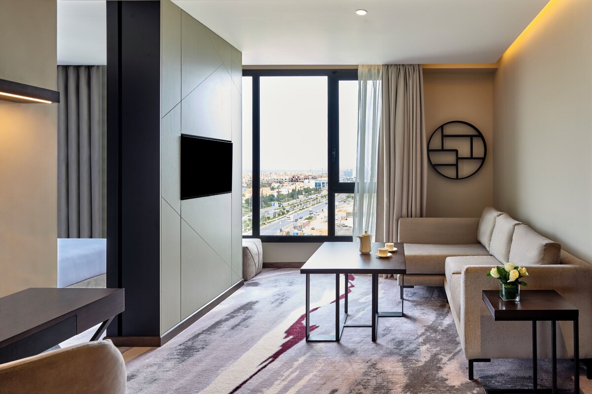 Une chambre d’hôtel moderne dotée d’une grande fenêtre, d’un téléviseur à écran plat, d’un canapé, d’une table basse et d’une petite table d’appoint sur laquelle est posé un vase de fleurs.