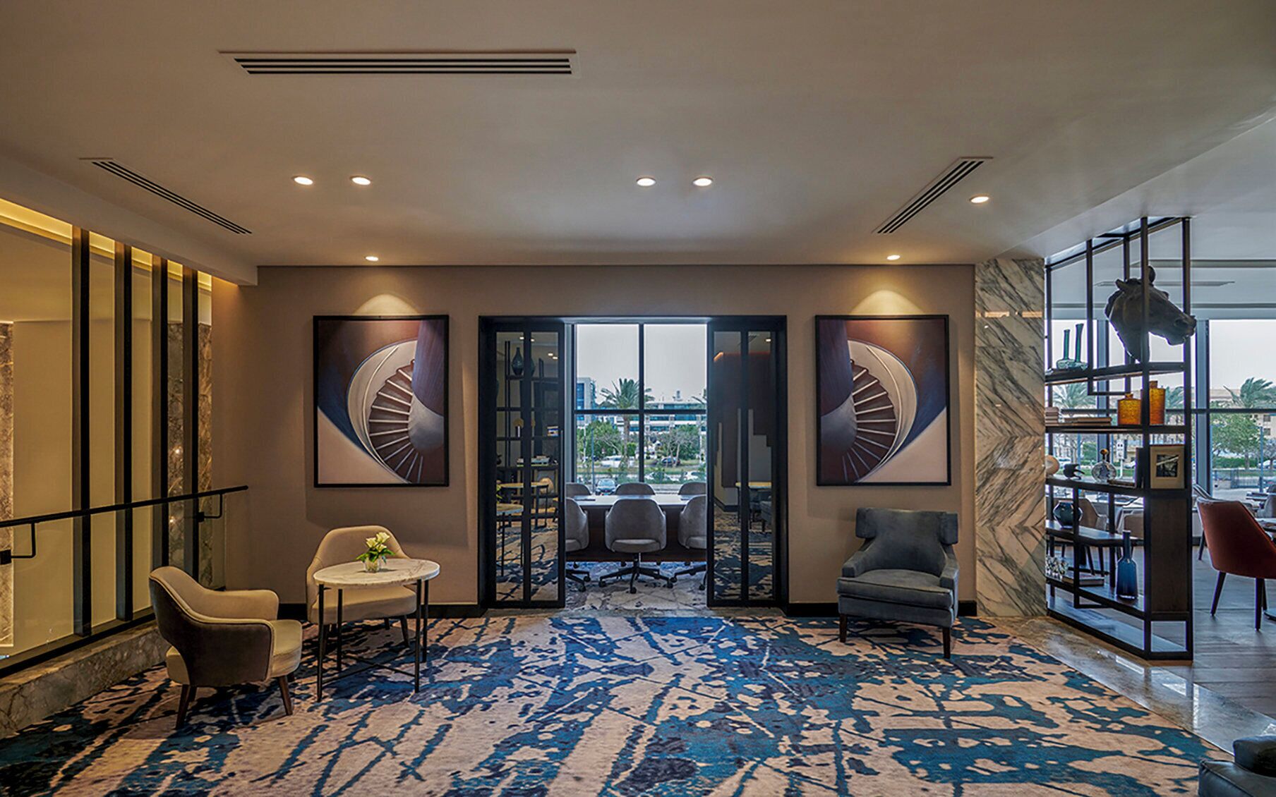 A modern lobby with a blue patterned carpet, a seating area with chairs and a table, and a large window with a view of palm trees and buildings.