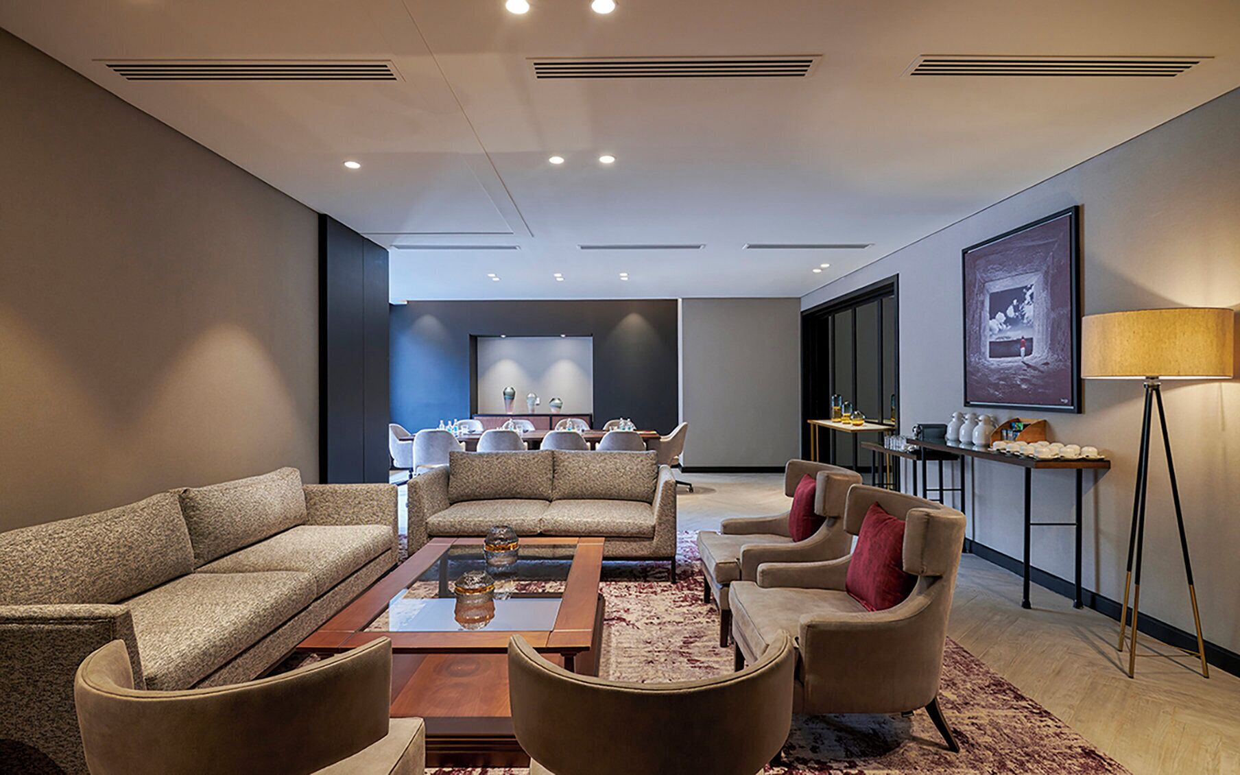 Una habitación de hotel moderna con un conjunto de sofá, una mesa de centro, un comedor con sillas, una mesita de noche con lámpara y una fotografía grande en el marco en la pared.