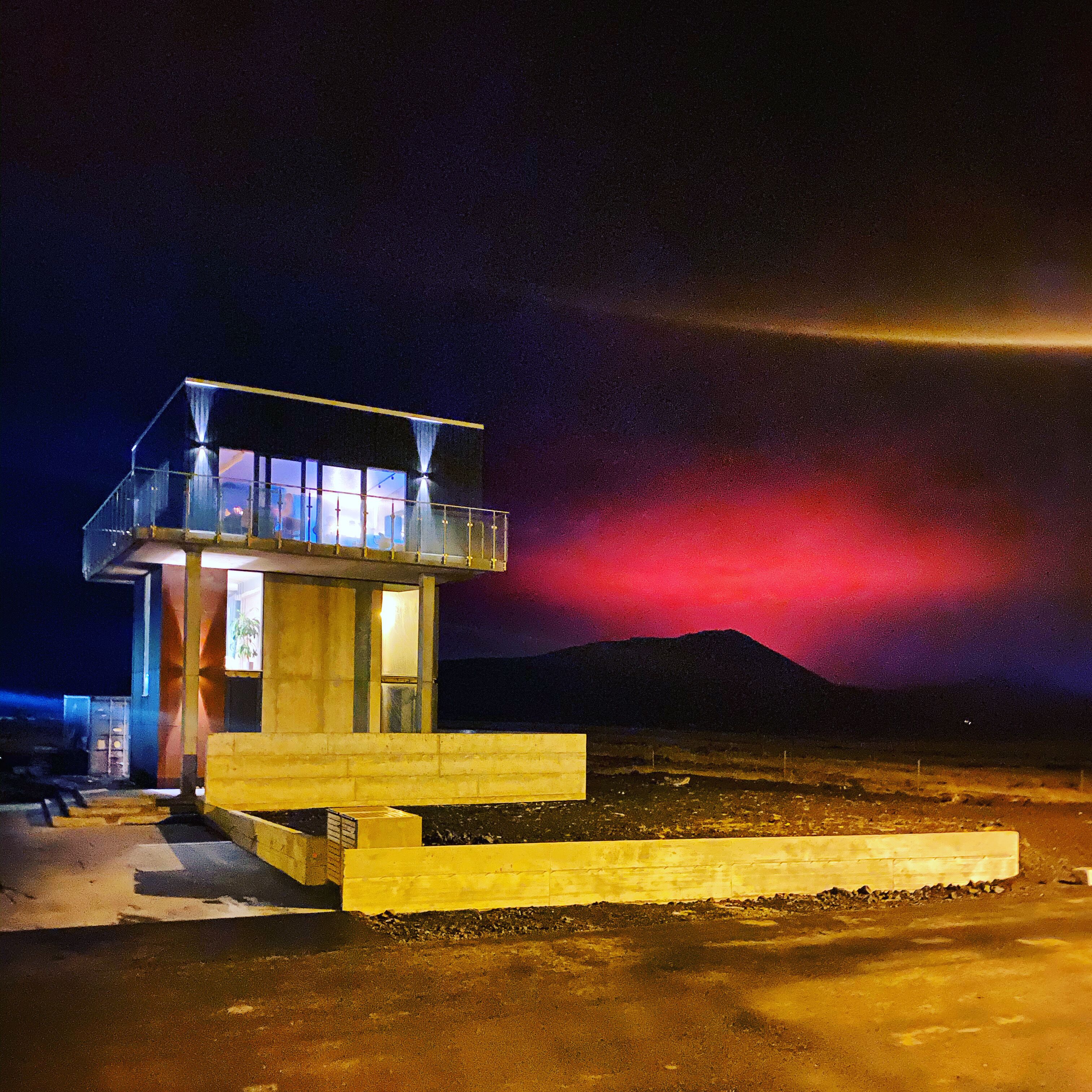 A small, single-story building with a balcony, located in a desert-like environment with a mountain in the background.