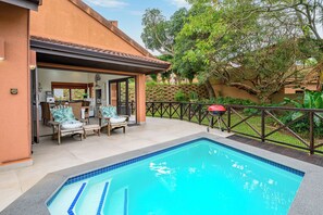 A swimming pool with a tiled deck, a covered patio area with outdoor furniture, and a view of a natural landscape.
