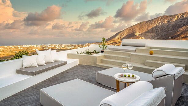 A rooftop terrace with a seating area, a table with glasses and fruit, and a view of a city and mountains.